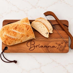 May include: A rectangular wooden bread board with a dark brown leather hanging loop, engraved with the name Brianna and a floral design. A wooden bread slicer with a metal wire is positioned on the board next to a loaf of bread and two slices.