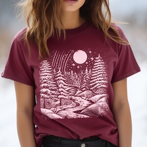 May include: A maroon t-shirt with a pink graphic print of a snowy forest scene with a full moon and shooting stars.