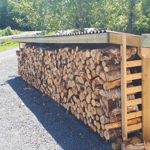 K&ouml;nnte beinhalten: Ein Holzscheiter-Schuppen mit einem Metalldach, gestapelt mit Holzscheiten. Der Schuppen hat eine Holzlattenwand auf der rechten Seite.