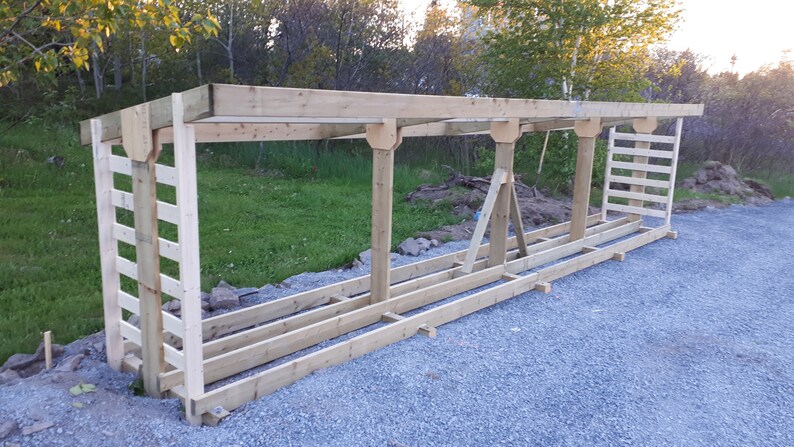 K&ouml;nnte beinhalten: Ein Holz-Brennholzregal mit Dach und einer Leiter an der Seite. Das Regal ist aus unbehandeltem Holz und steht auf einem Kiesweg.