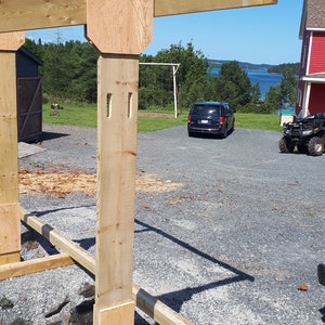 K&ouml;nnte beinhalten: Ein Holzrahmenbauprojekt mit einem Balken, der von einem Pfosten getragen wird. Der Pfosten ist mit einem St&uuml;ck Sperrholz an einem Holzsockel befestigt. Der Rahmen wird in einem Garten mit einem blauen Auto und einem roten Haus im Hintergrund gebaut.