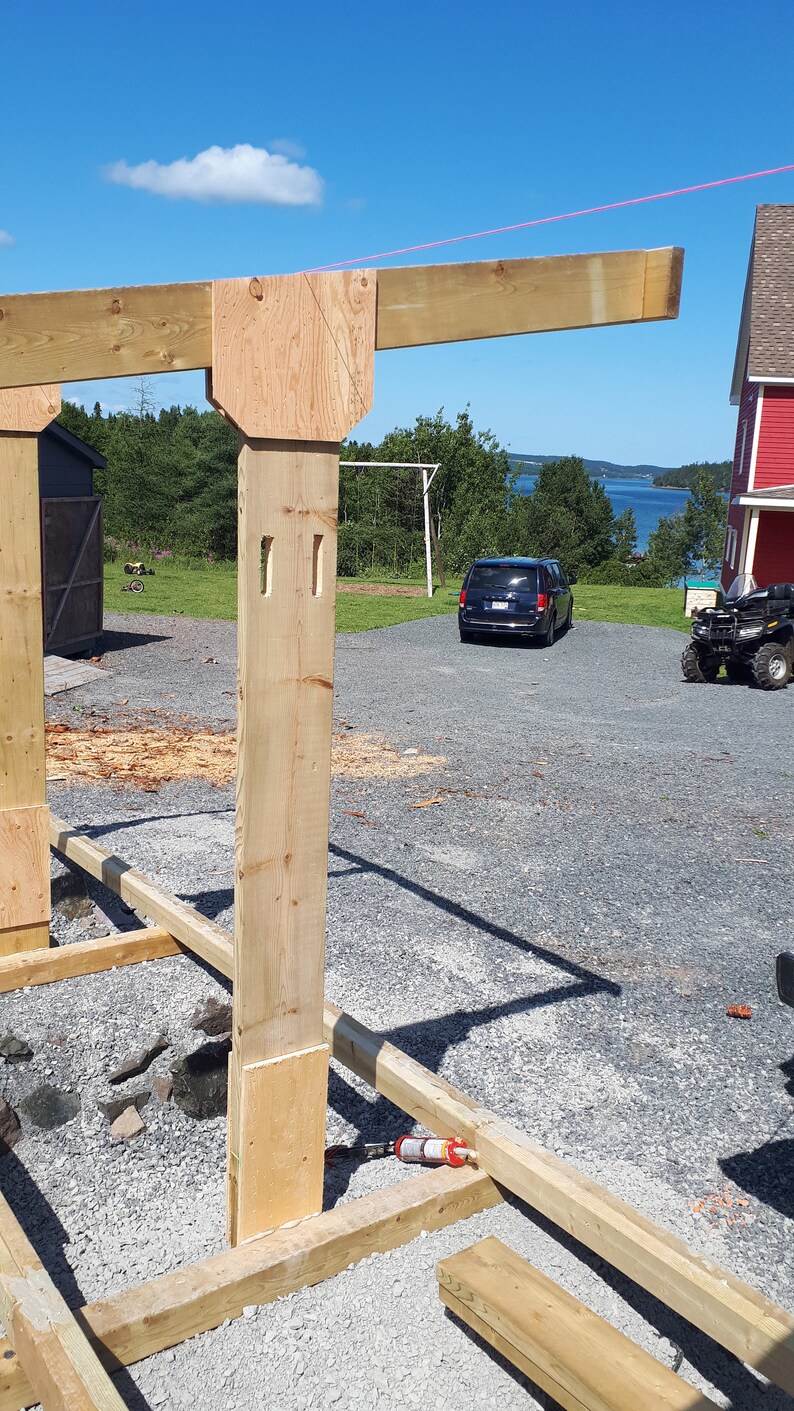 K&ouml;nnte beinhalten: Ein Holzrahmenbauprojekt mit einem Balken, der von einem Pfosten getragen wird. Der Pfosten ist mit einem St&uuml;ck Sperrholz an einem Holzsockel befestigt. Der Rahmen wird in einem Garten mit einem blauen Auto und einem roten Haus im Hintergrund gebaut.