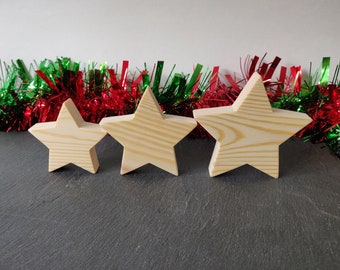 May include: Three wooden stars of different sizes, painted in a natural wood color, are arranged in a row on a black surface. The stars are in front of a red and green garland.