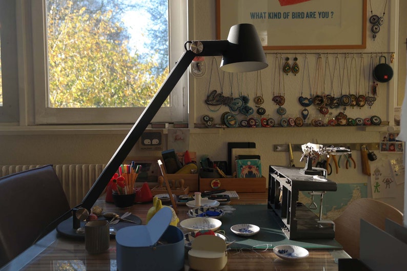 Puede incluir: Un espacio de trabajo con una l&aacute;mpara de escritorio, una mesa con diversos materiales de artesan&iacute;a y una pared decorada con collares coloridos y el texto "&iquest;Qu&eacute; tipo de p&aacute;jaro eres?"