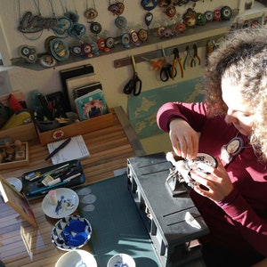 Puede incluir: Una persona trabajando en un proyecto artesanal en una mesa con varias herramientas y suministros. Est&aacute; sosteniendo un peque&ntilde;o objeto redondo blanco con dise&ntilde;os en blanco y negro. La mesa est&aacute; cubierta con una estera de corte verde y tiene una variedad de herramientas y suministros, incluyendo un porta l&aacute;pices rojo, una caja de madera con sellos y un peque&ntilde;o plato con un dise&ntilde;o azul y blanco.