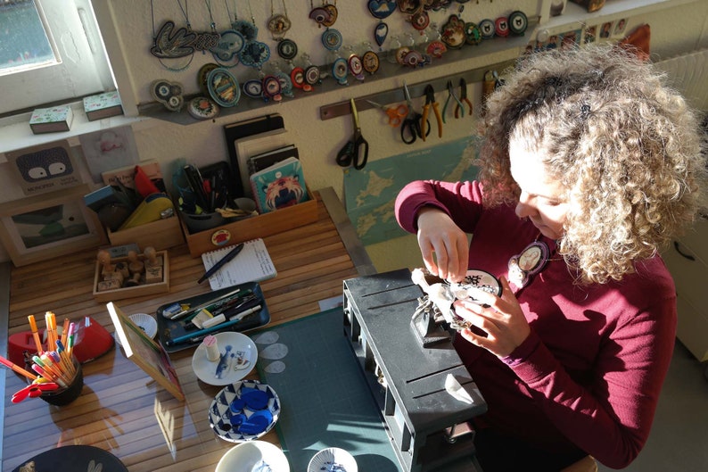 Puede incluir: Una persona trabajando en un proyecto artesanal en una mesa con varias herramientas y suministros. Est&aacute; sosteniendo un peque&ntilde;o objeto redondo blanco con dise&ntilde;os en blanco y negro. La mesa est&aacute; cubierta con una estera de corte verde y tiene una variedad de herramientas y suministros, incluyendo un porta l&aacute;pices rojo, una caja de madera con sellos y un peque&ntilde;o plato con un dise&ntilde;o azul y blanco.