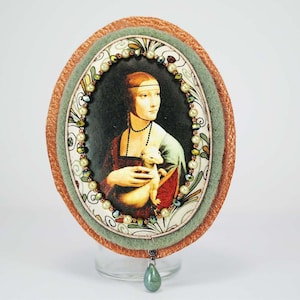 Puede incluir: Broche de forma ovalada con un marco de tela verde y naranja. El centro presenta una imagen impresa de una mujer sosteniendo un animal peque&ntilde;o. El marco est&aacute; decorado con perlas y cuentas.