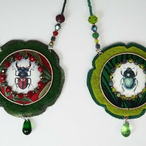Puede incluir: Dos collares de fieltro verde con colgantes redondos que presentan ilustraciones de escarabajos. Un colgante tiene un fondo rojo y verde con un escarabajo rojo, el otro tiene un fondo verde y negro con un escarabajo azul.