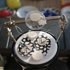 Puede incluir: Dos pendientes de tela en blanco y negro con cuentas azules, sostenidos en una abrazadera con c&iacute;rculos de fieltro blanco y un recipiente transparente de cuentas azules.