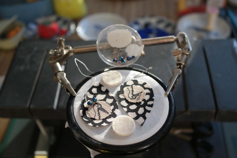 Puede incluir: Dos pendientes de tela en blanco y negro con cuentas azules, sostenidos en una abrazadera con c&iacute;rculos de fieltro blanco y un recipiente transparente de cuentas azules.