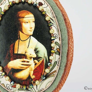 Puede incluir: Una obra de arte redonda, de forma ovalada, enmarcada con un borde verde y marr&oacute;n. La obra de arte presenta una pintura de una mujer sosteniendo un peque&ntilde;o animal blanco. La mujer lleva un vestido azul y rojo y un collar de perlas. El marco est&aacute; decorado con un borde de cuentas de perlas blancas y cuentas verdes y marrones.