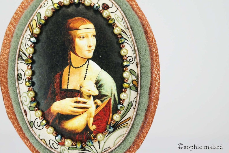 Puede incluir: Una obra de arte redonda, de forma ovalada, enmarcada con un borde verde y marr&oacute;n. La obra de arte presenta una pintura de una mujer sosteniendo un peque&ntilde;o animal blanco. La mujer lleva un vestido azul y rojo y un collar de perlas. El marco est&aacute; decorado con un borde de cuentas de perlas blancas y cuentas verdes y marrones.