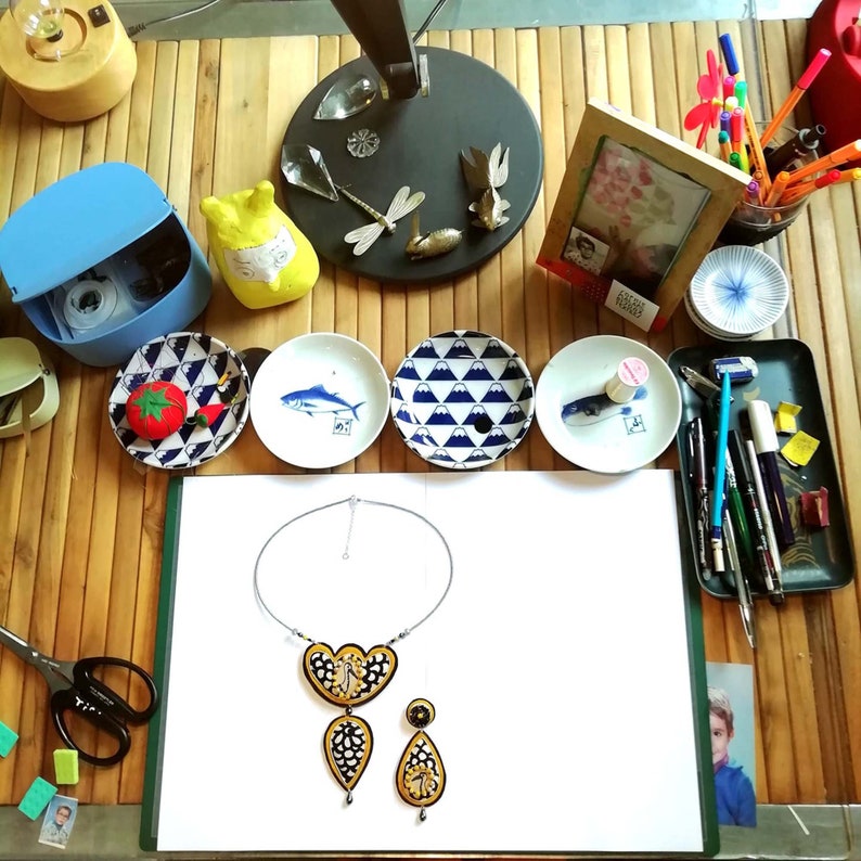 Puede incluir: Una hoja de papel blanca con un collar y pendientes plateados con un dise&ntilde;o negro y dorado. El collar tiene un colgante en forma de coraz&oacute;n con un dise&ntilde;o negro y dorado. Los pendientes son de forma de l&aacute;grima con un dise&ntilde;o negro y dorado. El papel est&aacute; sobre una mesa de madera con varios otros objetos, incluyendo una peque&ntilde;a caja azul, una peque&ntilde;a figurita amarilla y un peque&ntilde;o plato blanco con un dise&ntilde;o de pez.