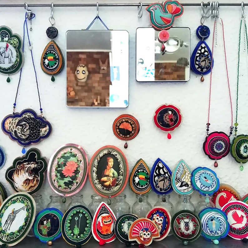 Puede incluir: Una colecci&oacute;n de joyas y art&iacute;culos decorativos hechos a mano, coloridos, que incluyen collares, aretes y broches. Los art&iacute;culos presentan una variedad de dise&ntilde;os, que incluyen animales, flores y patrones geom&eacute;tricos. Las joyas se exhiben en una pared blanca con espejos y otros elementos decorativos.