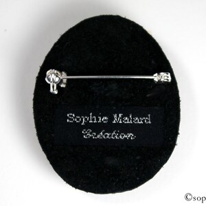 Puede incluir: Broche ovalada negra con un alfiler plateado y una etiqueta negra que dice "Sophie Malard Creation".