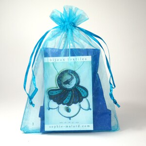 Puede incluir: Una bolsa de regalo de organza azul turquesa con un collar azul y negro en su interior. El collar presenta un colgante azul y negro con un dise&ntilde;o de ballena. La bolsa tiene una tarjeta blanca con el texto "bijoux textiles" y una direcci&oacute;n de sitio web.