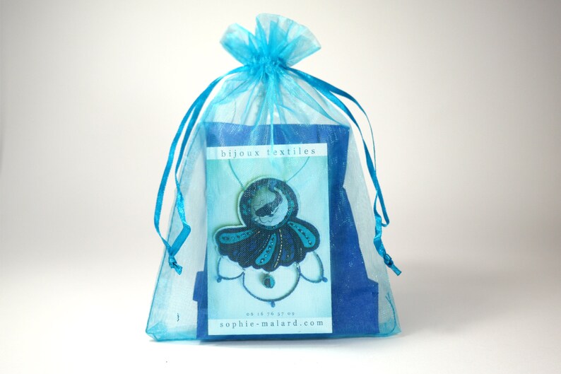 Puede incluir: Una bolsa de regalo de organza azul turquesa con un collar azul y negro en su interior. El collar presenta un colgante azul y negro con un dise&ntilde;o de ballena. La bolsa tiene una tarjeta blanca con el texto "bijoux textiles" y una direcci&oacute;n de sitio web.