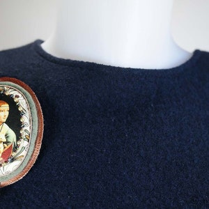 Puede incluir: Un fondo de tela azul oscuro con un broche ovalado marr&oacute;n que presenta una pintura de una mujer con un animal blanco. El broche tiene un colgante de piedra preciosa verde.