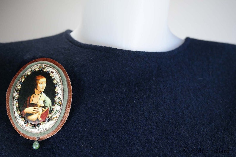 Puede incluir: Un fondo de tela azul oscuro con un broche ovalado marr&oacute;n que presenta una pintura de una mujer con un animal blanco. El broche tiene un colgante de piedra preciosa verde.