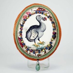 Peut inclure: Une broche ronde recouverte de tissu avec un motif floral coloré et une illustration bleue et blanche d'un dodo. La broche est entourée d'une bordure de perles et a un pendentif en pierre précieuse verte.