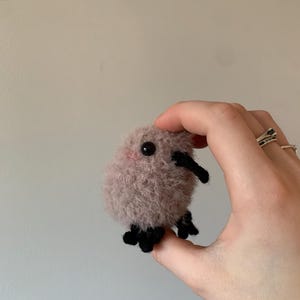 May include: A small, fuzzy, pink crocheted creature with black eyes and legs. The creature is being held in a hand.