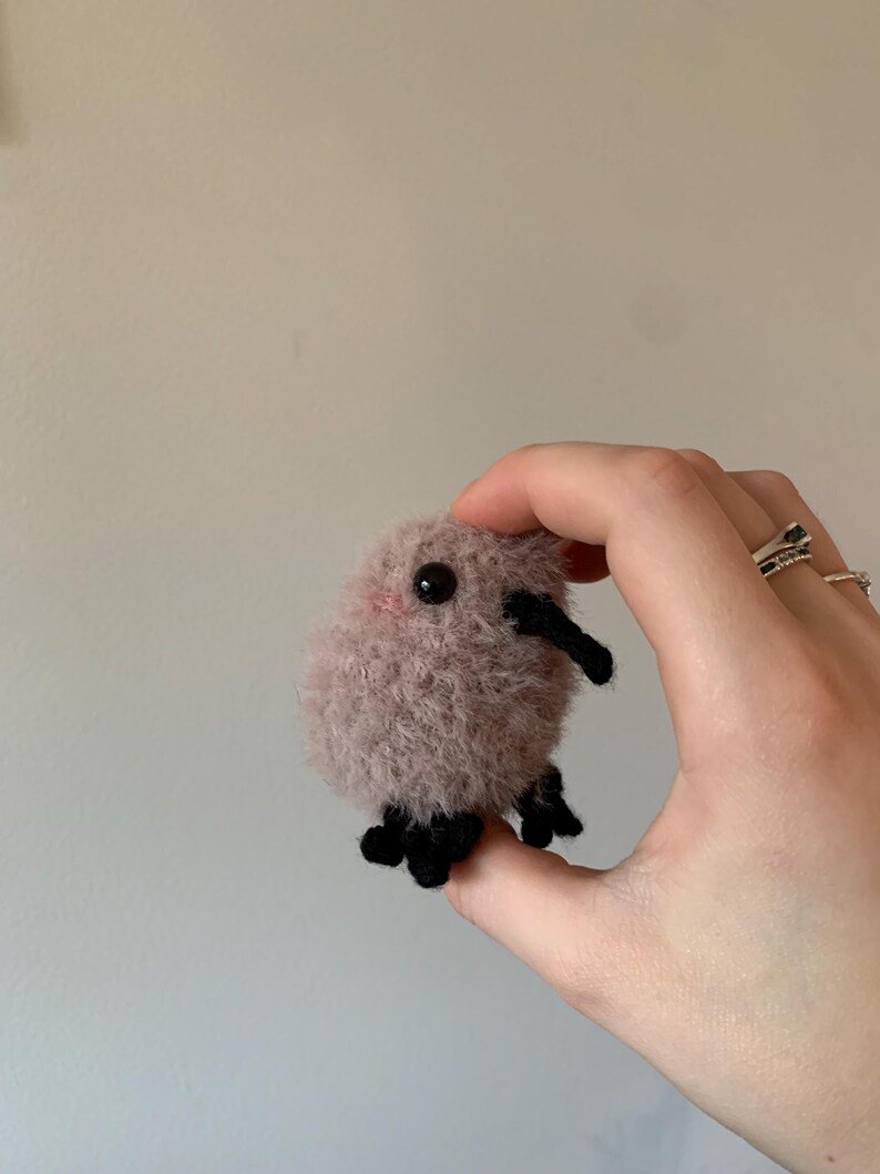 May include: A small, fuzzy, pink crocheted creature with black eyes and legs. The creature is being held in a hand.