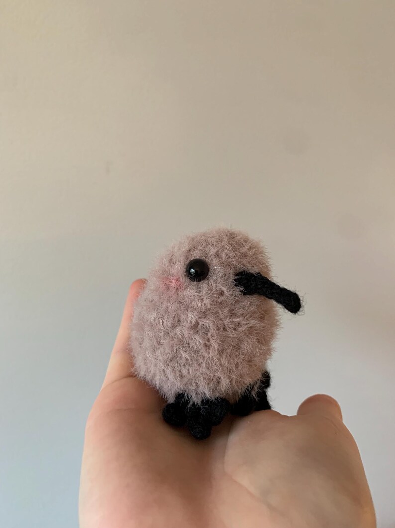 May include: A small, fluffy, crocheted stuffed animal in the shape of a bird. The bird is light brown with black eyes and a black beak. It has black crocheted legs.