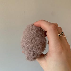 May include: A soft, fluffy, light pink makeup brush. The brush is held in a hand.