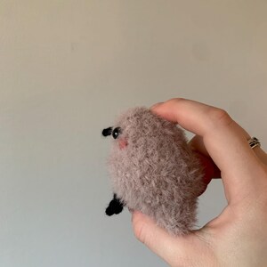May include: A small, fluffy, pink and brown plush toy with black eyes and a small black nose. The toy is being held in a hand.