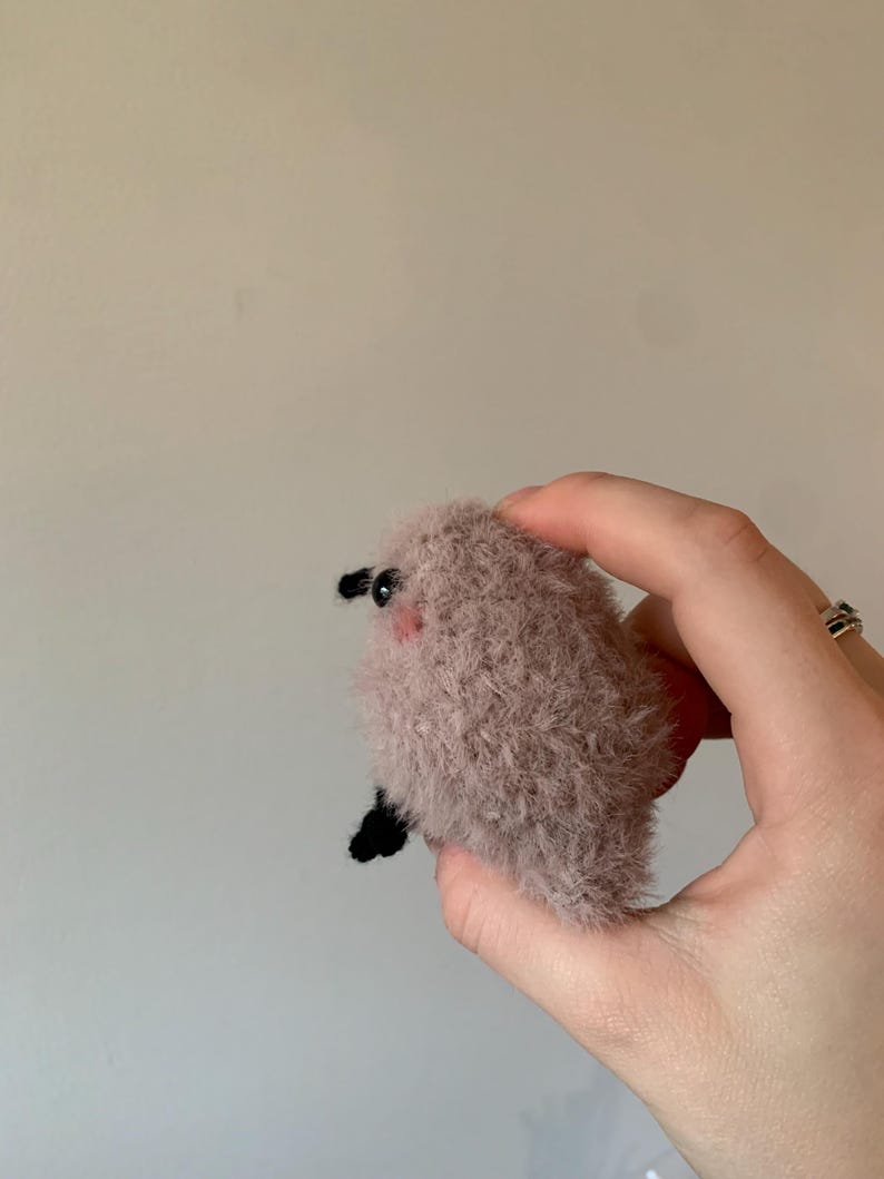 May include: A small, fluffy, pink and brown plush toy with black eyes and a small black nose. The toy is being held in a hand.