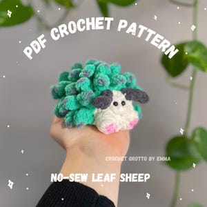 May include: A crochet plush toy shaped like a sheep with a textured green and purple body, a white face, dark gray ears, and pink feet. The image includes the text PDF CROCHET PATTERN, CROCHET GROTTO BY EMMA, and NO-SEW LEAF SHEEP.