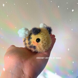 May include: A handmade, plush bee with yellow, black, and white accents. The bee has a round body, black eyes, and small white wings. The text "CROCHET GROTTO BY EMMA" is visible.