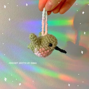 May include: A crocheted hummingbird ornament made with green, pink and white yarn. The ornament is hanging from a white and pink string.