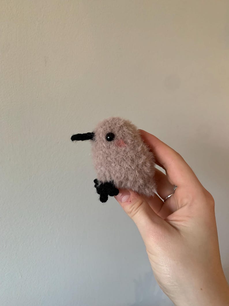 May include: A small, pink, fuzzy, crocheted bird with black eyes and a black beak. The bird is being held in a person's hand.