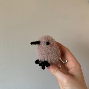 May include: A small, pink, fuzzy bird with black eyes and a black beak. The bird has black feet and is crocheted.