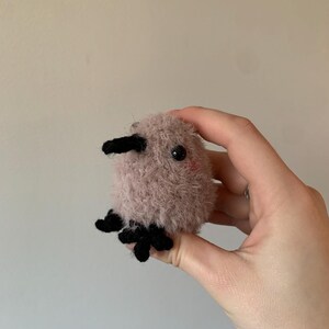 May include: A small, pink, fuzzy, crocheted stuffed animal with black eyes and a black beak. The animal has four black crocheted feet.