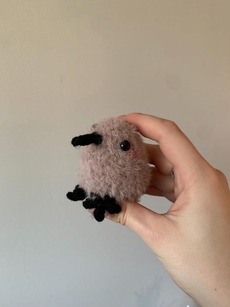 May include: A small, pink, fuzzy, crocheted stuffed animal with black eyes and a black beak. The animal has four black crocheted feet.