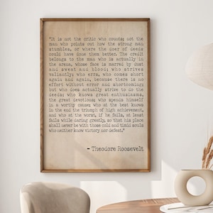 Könnte beinhalten: Ein gerahmter Druck mit einem Zitat von Theodore Roosevelt, der über einem Holztisch hängt. Das Zitat ist in schwarzem Text auf beigem Hintergrund. Der Raum umfasst auch eine beige Pendelleuchte und eine Vase mit getrockneten Pflanzen.