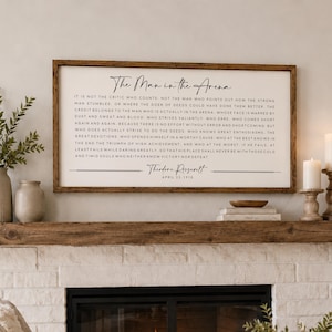 May include: A rectangular wooden framed wall sign featuring the text of The Man in the Arena speech by Theodore Roosevelt. The sign has a white background with black lettering and is mounted above a fireplace mantel with a natural wood finish.