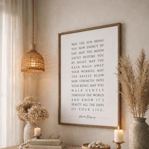 May include: A rectangular wall art print in a wooden frame featuring black text on a white background that reads: May the sun bring you new energy by day, may the moon softly restore you by night, may the rain wash away your worries.
