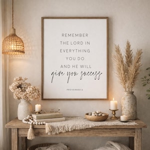 May include: A rectangular wall art print in a wooden frame featuring the black text: REMEMBER THE LORD IN EVERYTHING YOU DO. AND HE WILL give you success PROVERBS 3:6. The print is displayed on a wall above a wooden console table with decor.