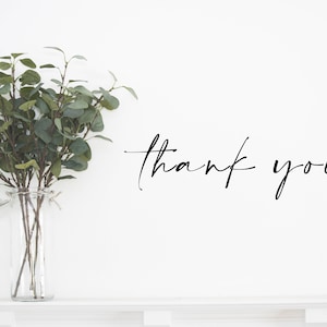 May include: A clear glass vase holds green eucalyptus branches. The words "thank you" are written in elegant black script on a white background. The vase sits on a white shelf.
