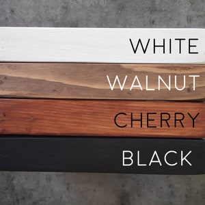 May include: Four wooden boards stacked on top of each other, showing different wood stains. The top board is white, the second board is walnut, the third board is cherry, and the bottom board is black.