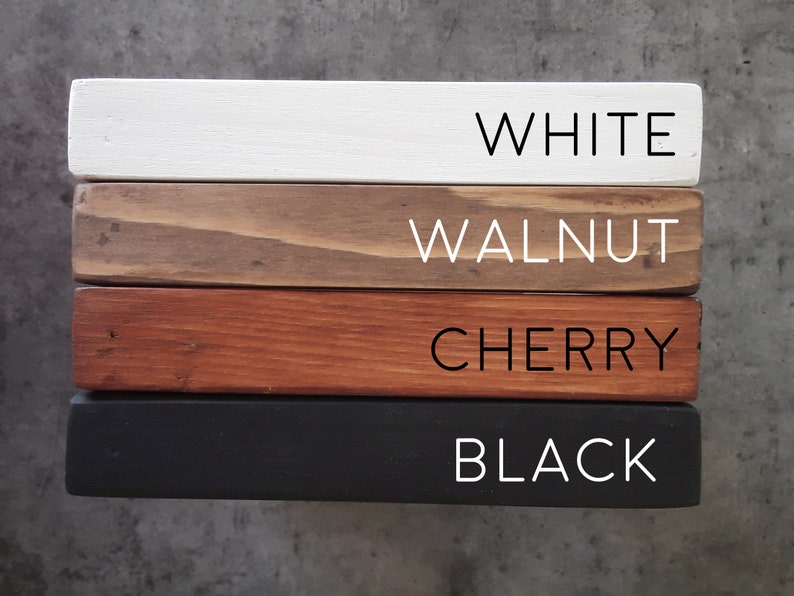 May include: Four wooden boards stacked on top of each other, showing different wood stains. The top board is white, the second board is walnut, the third board is cherry, and the bottom board is black.