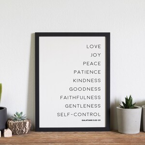 May include: A framed print with a black border lists virtues in black text: Love, Joy, Peace, Patience, Kindness, Goodness, Faithfulness, Gentleness, and Self-Control. The text "Galatians 5:22-23" is at the bottom. Small potted plants and decorative houses are on a wooden surface.