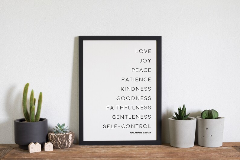 May include: A framed print with a black border lists virtues in black text: Love, Joy, Peace, Patience, Kindness, Goodness, Faithfulness, Gentleness, and Self-Control. The text "Galatians 5:22-23" is at the bottom. Small potted plants and decorative houses are on a wooden surface.