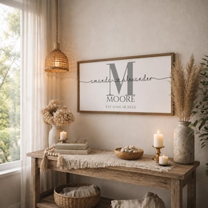 May include: A rectangular wall sign with a wooden frame featuring a large gray letter M, the names amanda and alexander connected by a heart, the surname MOORE, and the text EST JUNE 18, 2022 printed in black on a white background.