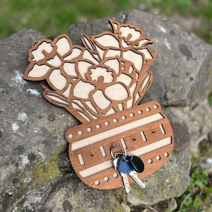 May include: Wooden key holder featuring a daffodil design. The key holder is made of light and dark brown wood, with a vase-shaped base and key hooks. A set of keys hangs from the hooks. The background is a blurred outdoor scene.