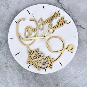May include: A white round clock with black minute markers and gold-colored hands. The clock features a gold stethoscope design, the text "Dr. Joanna Smith", and a floral accent. A decorative item for a doctor's office.