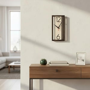Op de afbeelding: Een rechthoekige wandklok met een donkerbruin frame, zwarte cijfers op 12 en 6 en zwarte wijzers. De klok hangt aan een witte muur. Een houten console eronder bevat een kleine plant, boeken en een ingelijste foto.